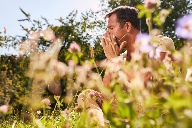 Man with hay fever allergy sits between blooming flowers and blows his nose - Vorteilhaft Leben - Vorteilhaft Leben