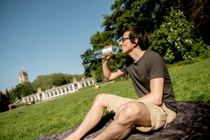 Mann sitzt auf einer Picknickdecke und trinkt aus einer Wasserflasche.
