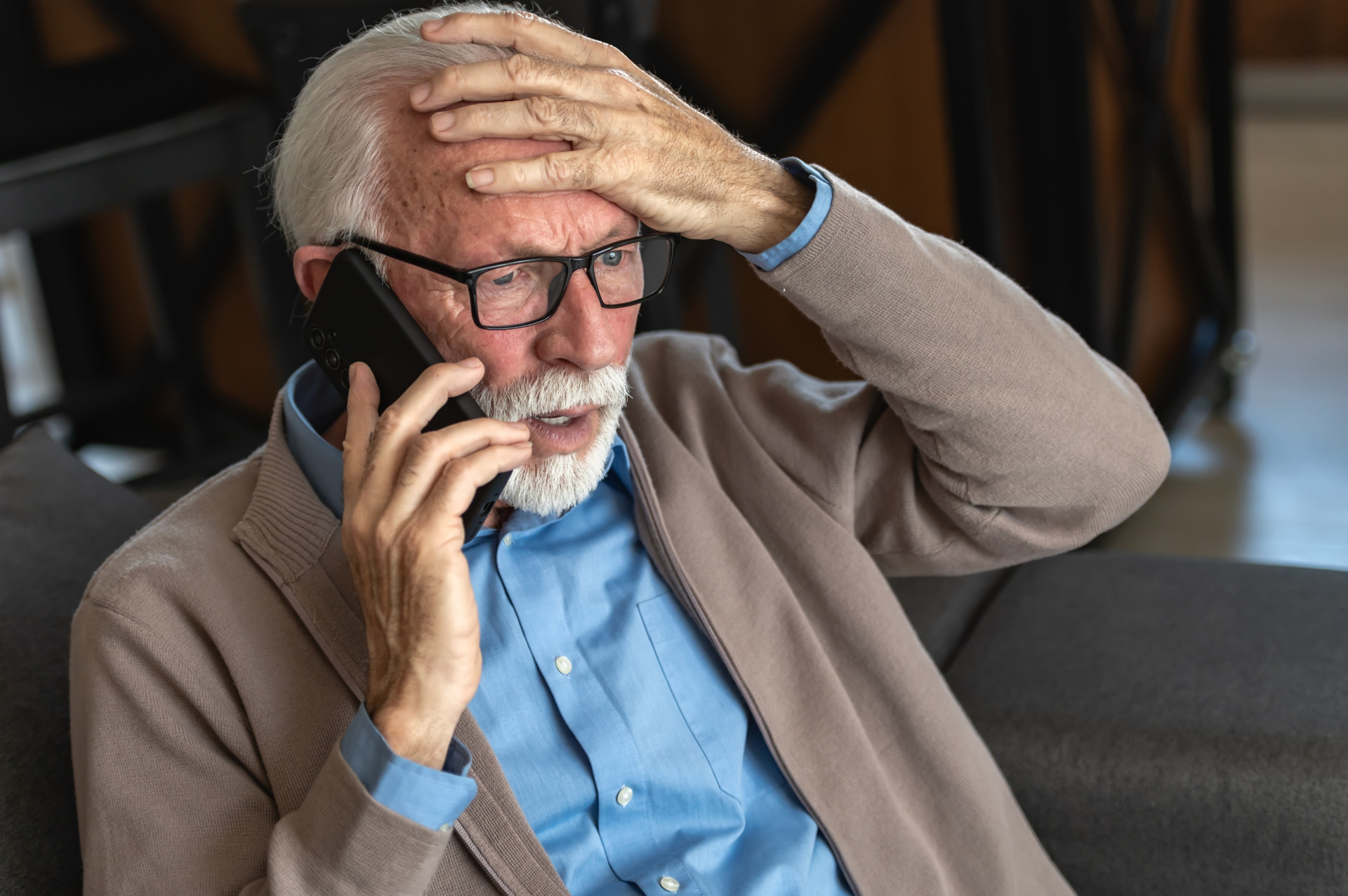 An elderly man with glasses and a beard wearing a blue shirt and beige cardigan looks distressed while talking on the phone holding his head in concern - Vorteilhaft Leben - Vorteilhaft Leben